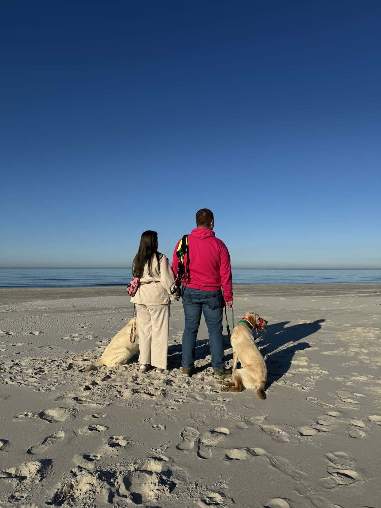 Para ludzi stojących tyłem na plaży przy spokojnym morzu.  
Po lewej stronie stoi kobieta w jasnym, beżowym dresie z długimi, ciemnymi włosami. Na plecach ma mały różowo-czarny plecak.  
Obok niej, po prawej, stoi mężczyzna w ciemnych dżinsach i jaskrawej, różowej lub czerwonej bluzy z kapturem. Na ramieniu ma ciemny plecak z kolorowymi paskami.  
Przed nimi rozciąga się szeroka, piaszczysta plaża i spokojna tafla wody, a nad nią jednolite, bezchmurne, intensywnie niebieskie niebo.  
Przy każdej z osób jest po jednym psie w jasnym, kremowym kolorze, przypominającym labradory lub podobną rasę. Pies po lewej leży na piasku, częściowo obrócony bokiem do aparatu. Pies po prawej siedzi obok mężczyzny, z założoną na kufę czerwoną obrożą lub kagańcem i zieloną bandamą lub obrożą na szyi.  
Piasek jest jasny, z licznymi śladami ludzkich stóp. Światło jest mocne, słoneczne, cienie osób i psów padają długie w prawą stronę, co sugeruje wczesny ranek lub późne popołudnie.  
Cała scena sprawia spokojne, spacerowe wrażenie: dwie osoby stoją obok siebie, trzymając smycze psów i patrząc w kierunku morza.